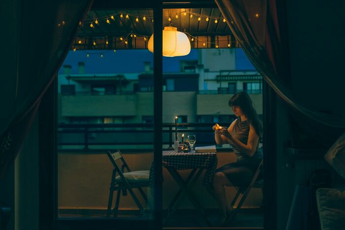 Young woman alone at a dimly lit table on a balcony in the evening, reflecting on family and golden child stories.