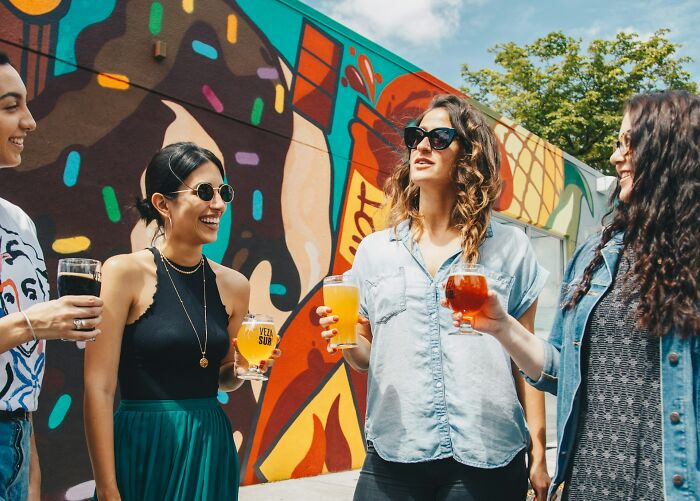 Group of women socializing outdoors, holding drinks near a colorful mural, representing subtle signs of cheating unnoticed.