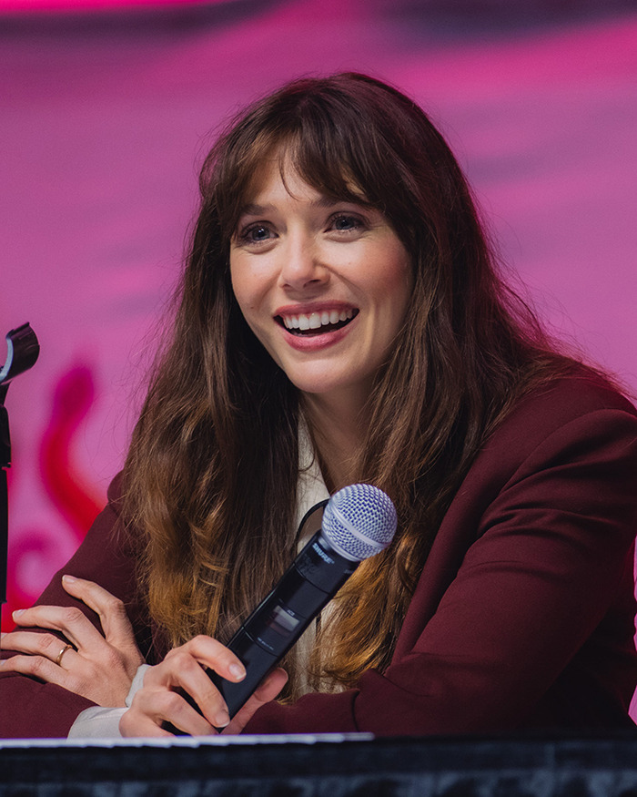 Elizabeth Olsen speaking at a panel, holding a microphone, breaking down the viral 6,7 meme with a smile.