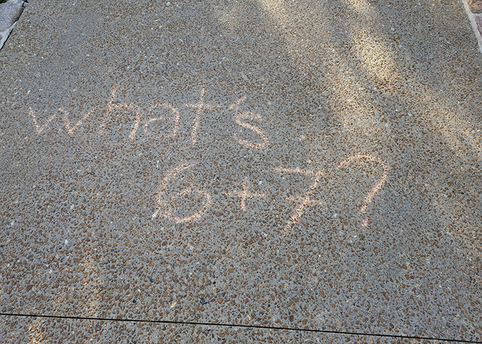Chalk writing on pavement asking what 6 plus 7 is, referencing the viral 6,7 meme involving Elizabeth Olsen.