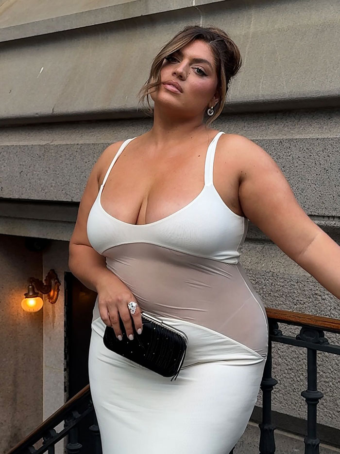 Plus-size model posing confidently in a white dress holding a black clutch against a stone wall backdrop. Plus-size model posing confidently in a white dress holding a black clutch against a stone wall backdrop.