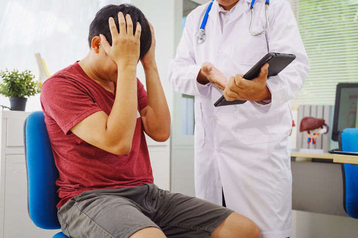 Paciente avergonzado en consulta médica, doctor con bata blanca usando tableta, experiencia incómoda en visita al doctor.