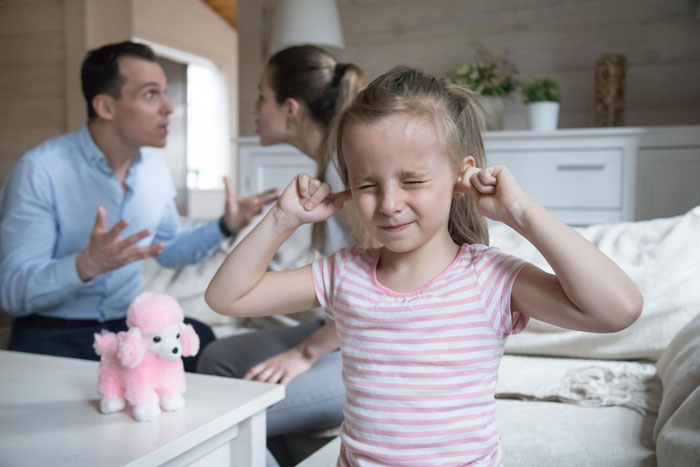 Young girl plugging ears as toxic dad and mom argue in living room, highlighting toxic family conflict and emotional distress.