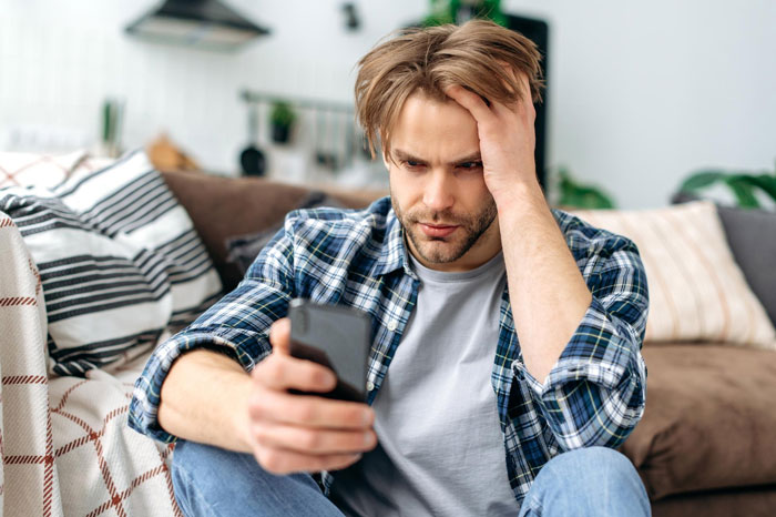 Man sitting on couch looking stressed while holding phone, reflecting on refusing to visit terminally ill ex-GF who cheated.
