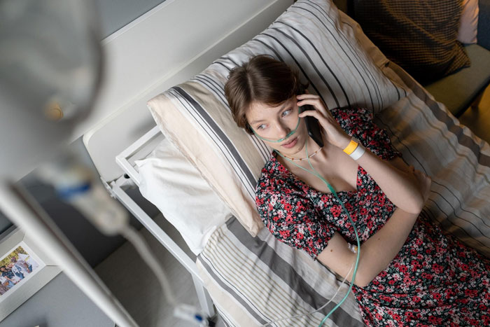 Young woman with oxygen tube lying in hospital bed talking on phone, illustrating terminally ill patient scenario.
