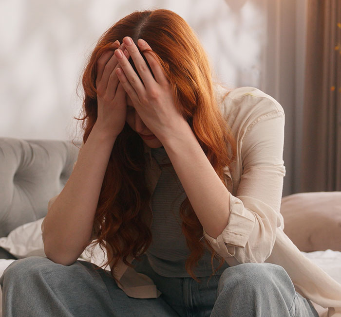 Red-haired woman sitting on a bed with her head in her hands, showing emotional distress and frustration. Red-haired woman sitting on a bed with her head in her hands, showing emotional distress and frustration.