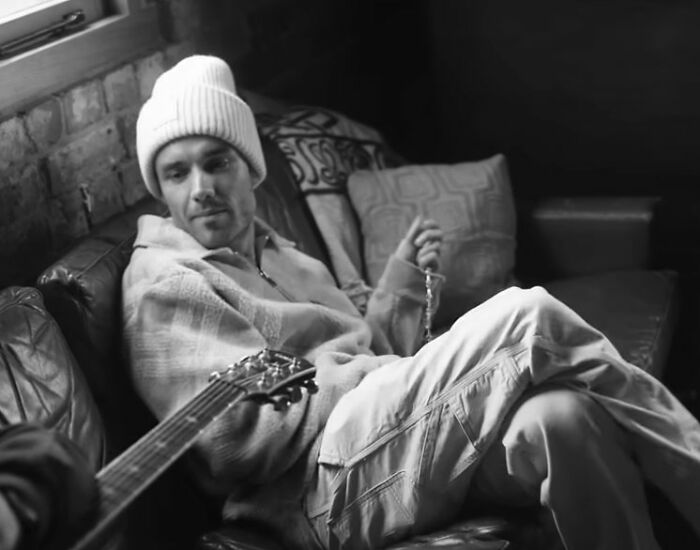 Man in a beanie and casual clothing sitting on a leather couch, holding a string of beads, related to Simon Cowell and Liam Payne.