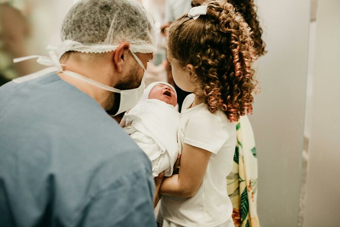 Father in surgical scrubs and mask showing newborn baby to young girl, sharing true facts that make others question reality.