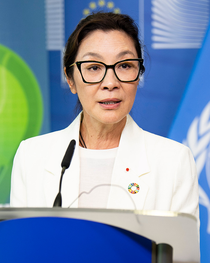 Michelle Yeoh speaking at a podium, wearing glasses and a white blazer, with a blue background in a formal setting. Michelle Yeoh speaking at a podium, wearing glasses and a white blazer, with a blue background in a formal setting.
