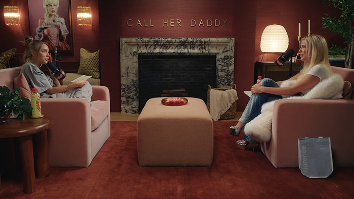 Two women sit on pink couches in a podcast studio discussing Millie Bobby Brown dating Jake Bongiovi with microphones.