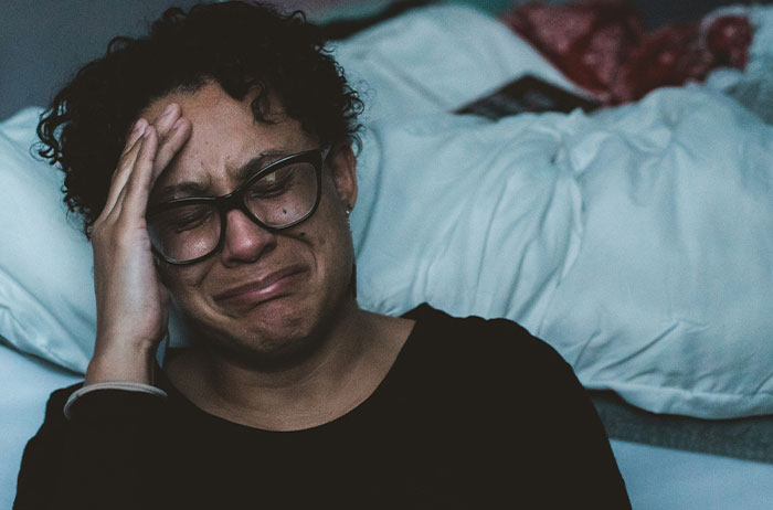 Woman with glasses crying and holding her head, expressing grief in a dimly lit room about unexpected lives ending.