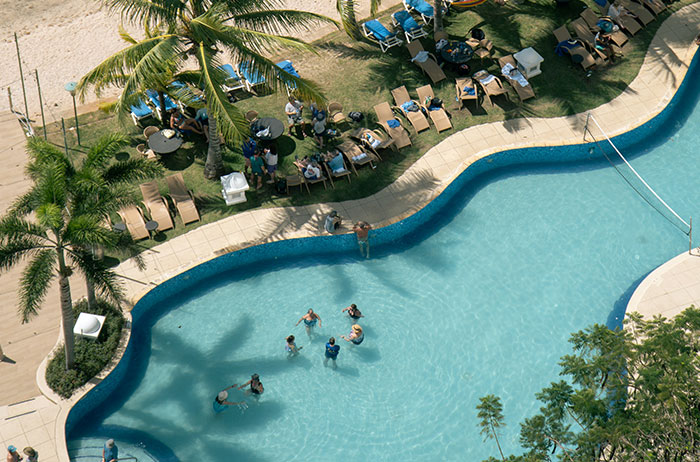 Aerial view of people swimming and relaxing by a pool surrounded by palm trees and lounge chairs at a resort.