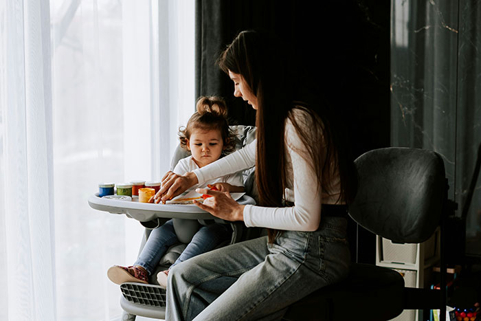 Woman using intuition while painting with her child in a high chair near a window with natural light.