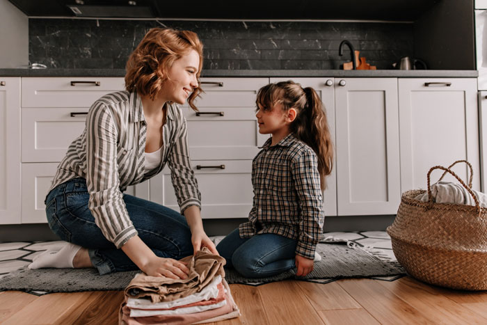 Young woman babysitting daughter in kitchen, folding clothes together, bonding during a calm home moment.