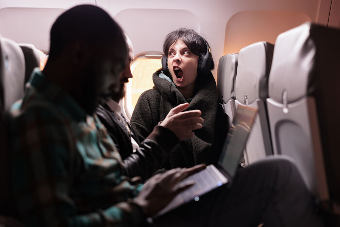Passenger yawning loudly wearing headphones on an airplane while flight crew work nearby on laptops during flight horror stories.
