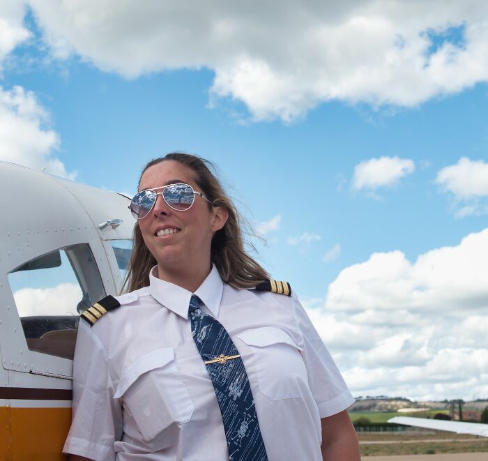 Female flight crew member wearing aviator sunglasses and uniform standing beside a small aircraft on a sunny day.