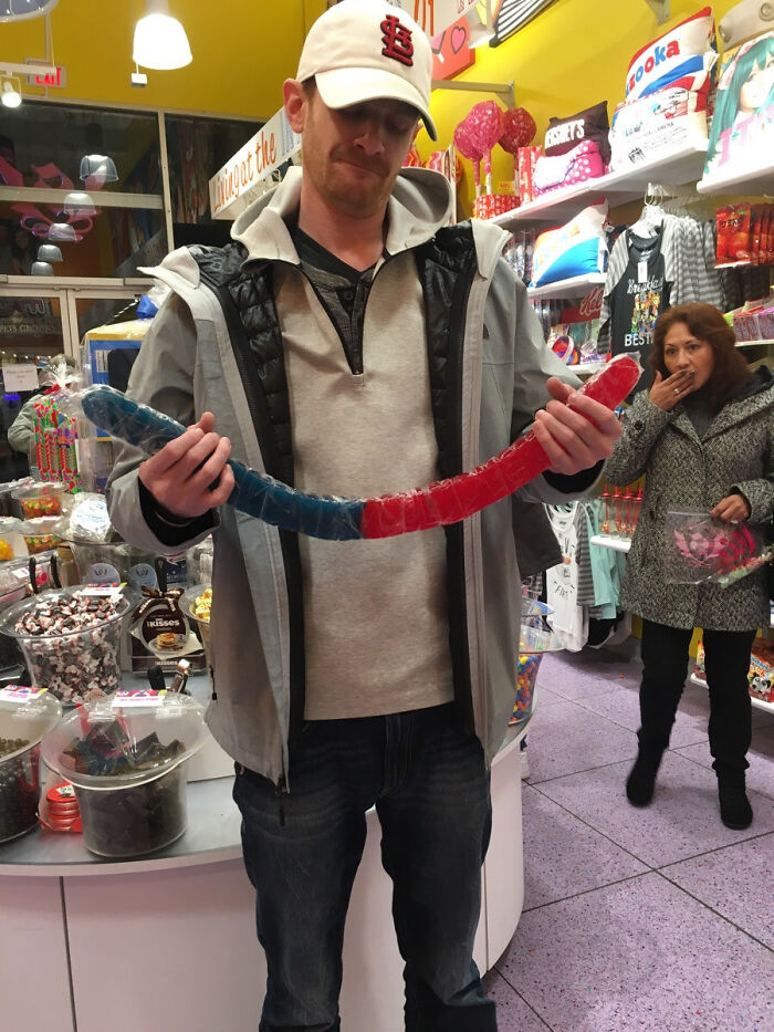 Man holding large red and blue candy in a store while a woman in the background reacts in a photobomb that stole the show.