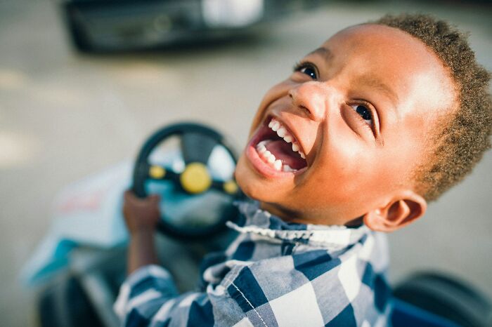 Young boy laughing while driving a toy car, showcasing kids doing something weird clearly passed down from parents.