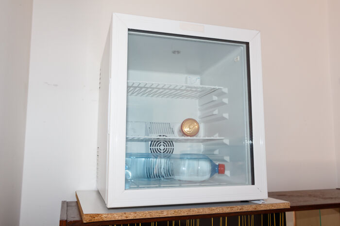Small white fridge with broken shelves and a single water bottle seen through the glass door inside a sparse room.