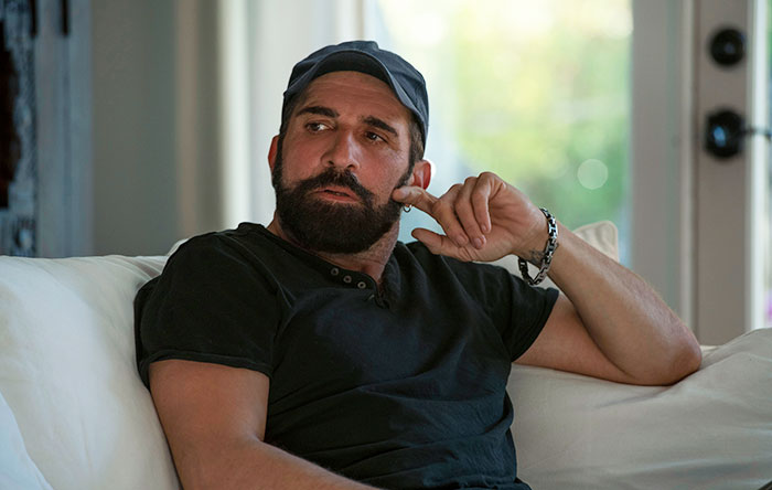 Man with beard and cap sitting on a couch looking away thoughtfully, representing a friend who doesn&rsquo;t understand clingy children.