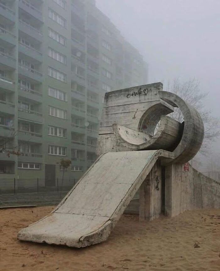Concrete playground slide with unusual unesthetic design in front of a grey, foggy residential building, highlighting ugly architecture.