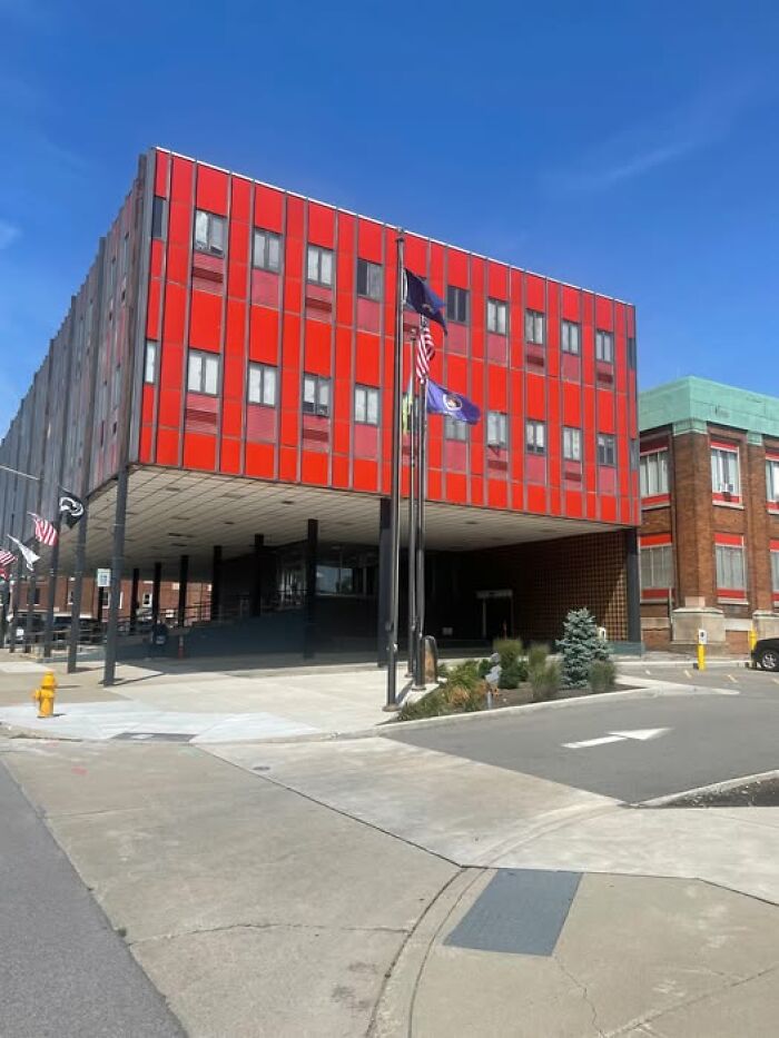Red and gray unesthetic building with flagpoles in front, showcasing ugly modern architecture on a clear day.
