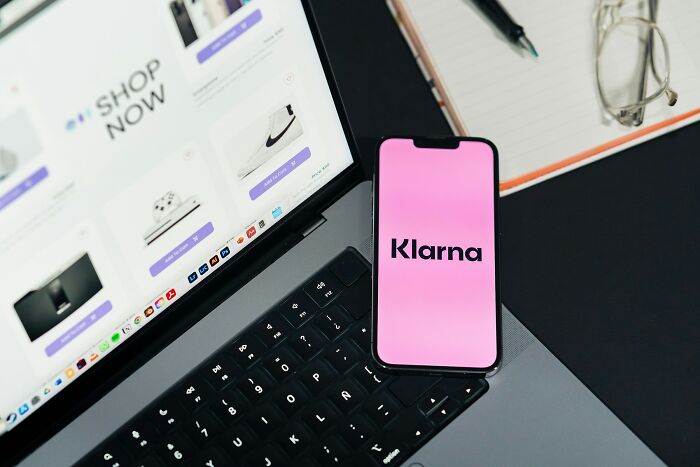 Smartphone displaying Klarna app on laptop keyboard with online shopping website and glasses nearby, highlighting online thread events.