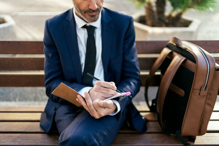 Man in blue suit writing notes on bench near leather backpack, illustrating etymology of common words concept.