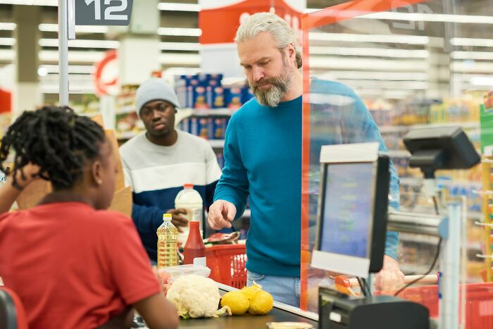 Man at grocery checkout counter looking concerned while cashier scans items, illustrating people falling for scams impact.