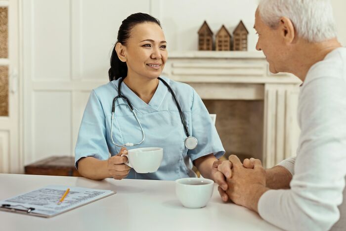 Nurse in scrubs with stethoscope talking to elderly man over coffee, illustrating family golden child stories concept.