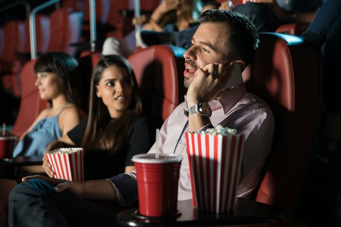 Man talking on phone in movie theater, ignoring companions showing toxic relationship traits with distracting behavior.