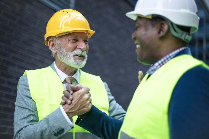 Two construction workers wearing safety helmets and vests shaking hands, illustrating work secrets and teamwork.