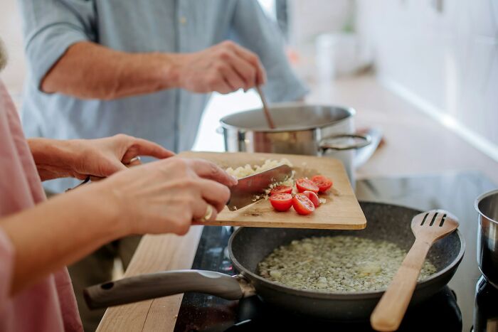 Two people cooking together, one chopping tomatoes and garlic with sharp knives on a wooden board.