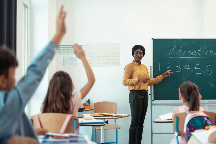 Teacher in classroom pointing at chalkboard with numbers while students raise hands showing work secrets in service jobs.