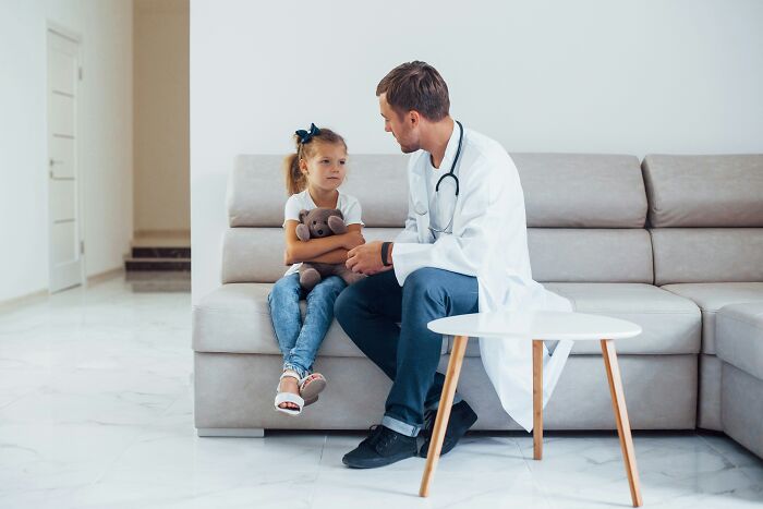 Doctor in white coat talking to a young girl patient holding a stuffed bear in a modern clinic setting.