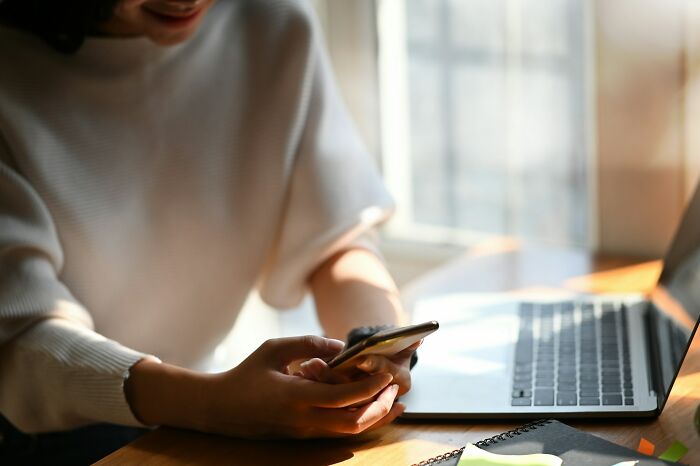 Person holding a smartphone near a laptop in soft natural light, depicting subtle signs of cheating behavior.
