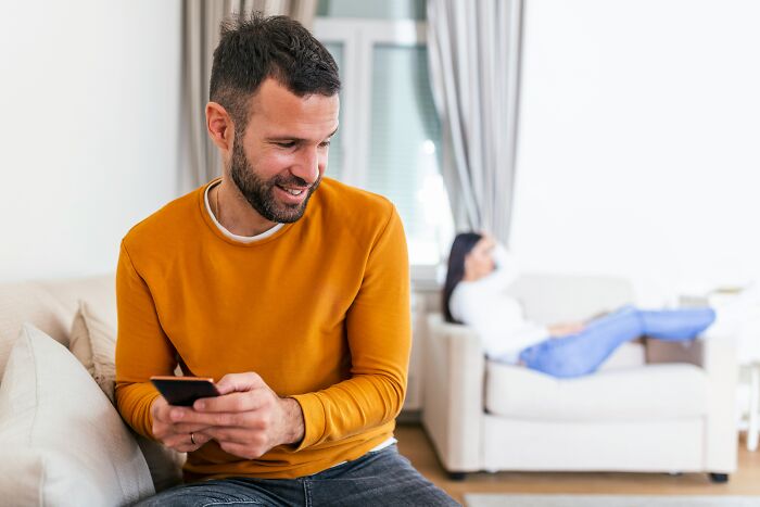 Man in an orange sweater on his phone showing signs of toxic relationship traits while woman sits upset in the background