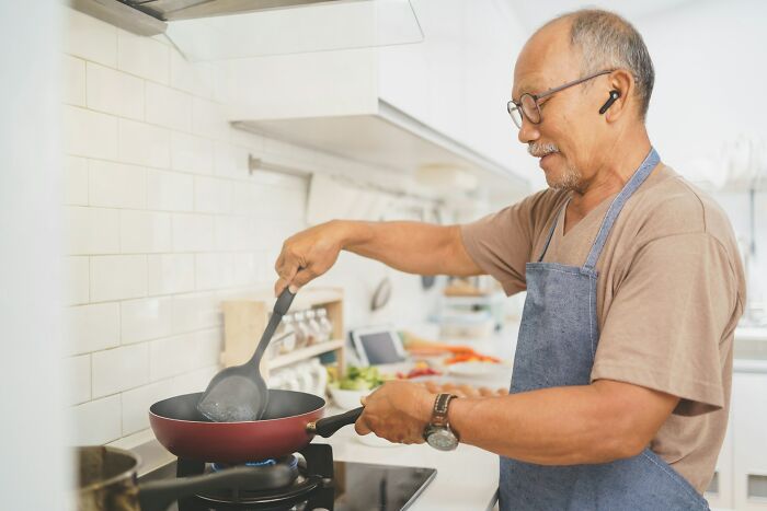 Older man wearing apron cooking on stove using spatula, demonstrating common home cooks mistakes with sharpen your knives advice.