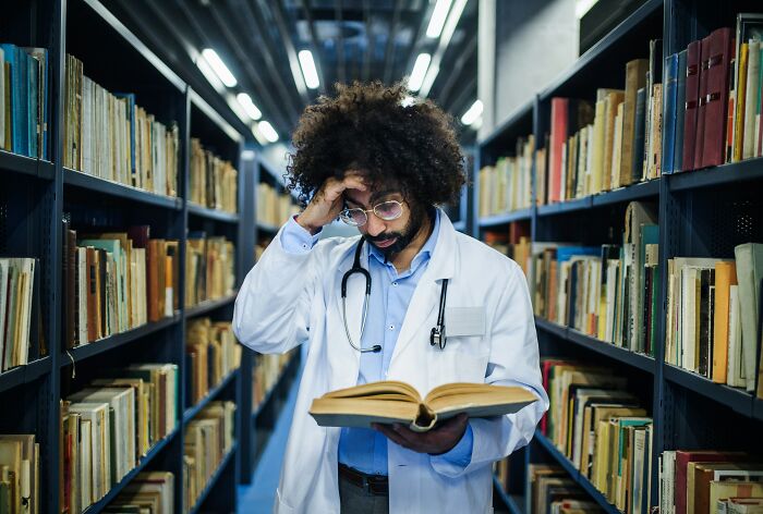 Man in white coat with stethoscope reading a book in a library, illustrating insider experiences of nepo babies.