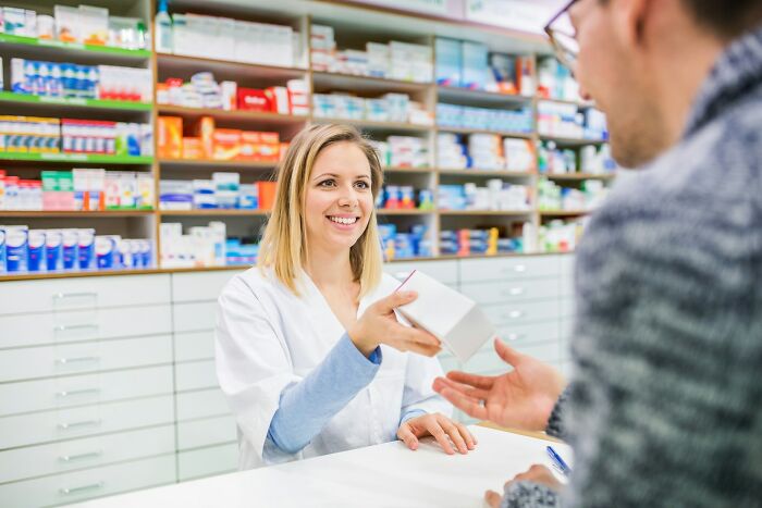 Pharmacist handing a medication box to a customer in a brightly lit pharmacy, illustrating trust in what often seems like a scam.
