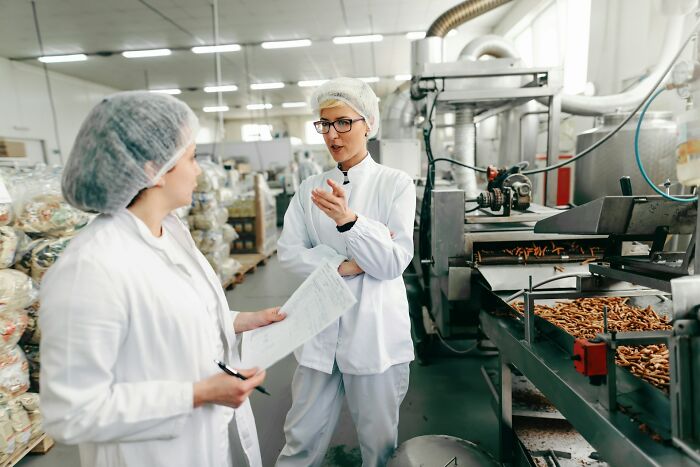 Two food factory workers wearing hairnets discussing production with machinery and trays of snacks in the background.