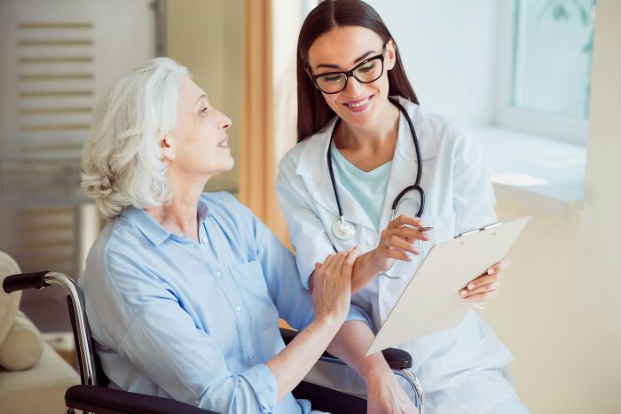 Female doctor with stethoscope smiling and holding clipboard while talking to elderly patient in wheelchair in clinic.