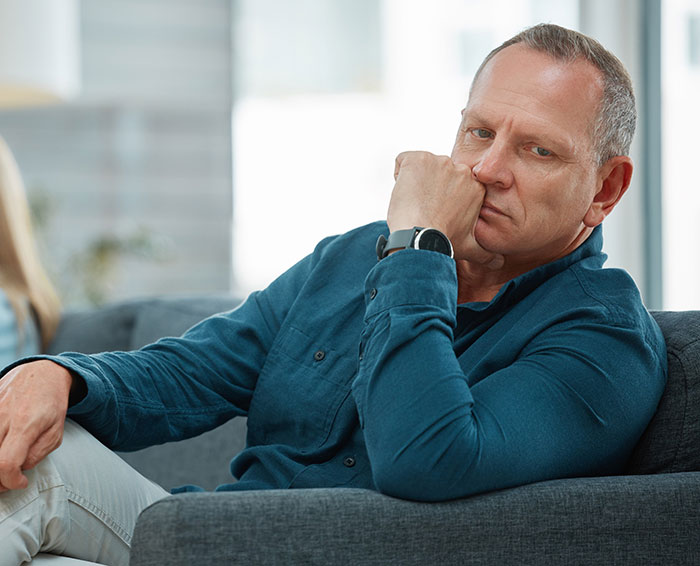 Middle-aged man sitting on a couch looking thoughtful and slightly frustrated while resting his fist against his face Middle-aged man sitting on a couch looking thoughtful and slightly frustrated while resting his fist against his face
