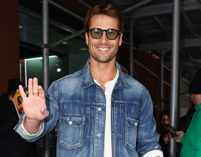 Glenn Powell smiling and waving, wearing sunglasses and a denim jacket, sharing emotional reason for flying UPS driver to SNL.