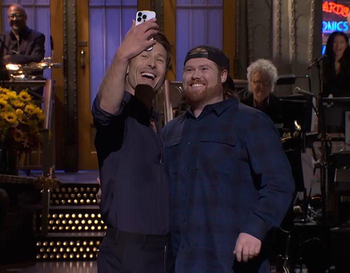 Glenn Powell smiling and taking a selfie with a UPS driver on stage during an emotional SNL moment.