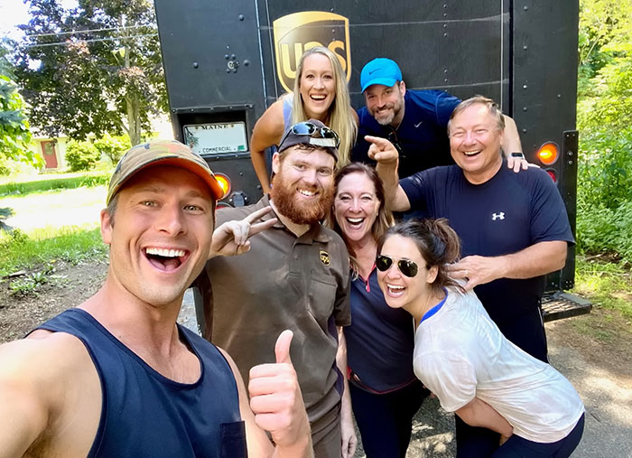 Glenn Powell with group smiling in front of UPS truck, celebrating flying UPS driver to SNL event outdoors.