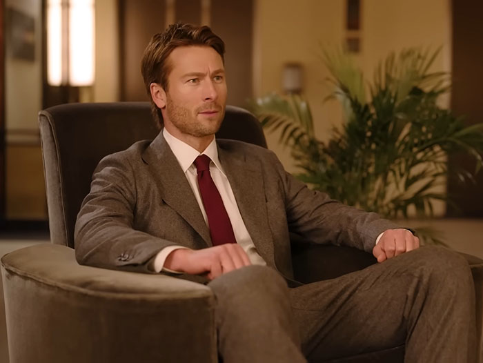 Glenn Powell seated in a suit, looking thoughtful, revealing emotional reason behind flying UPS driver to SNL.