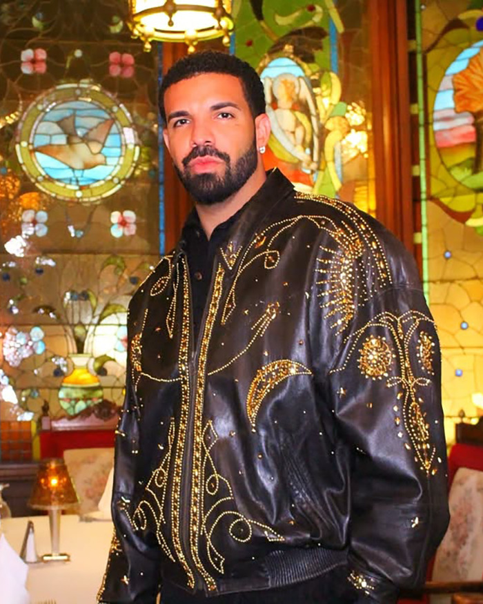 Drake wearing an embellished black leather jacket standing in front of stained glass windows at an indoor venue.