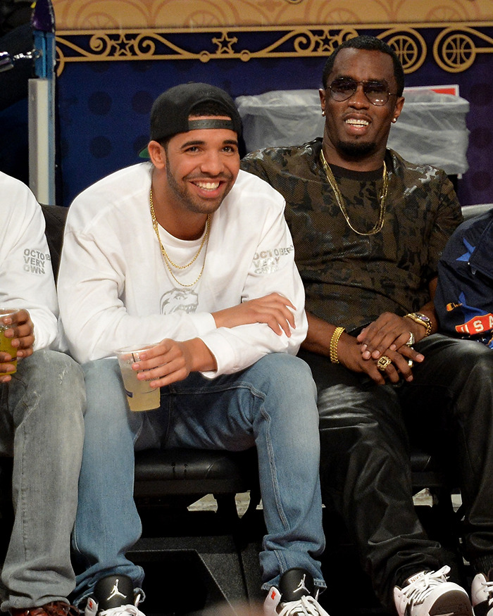 Drake sitting and smiling at an event, wearing a white sweatshirt and black cap, holding a drink in his hand.