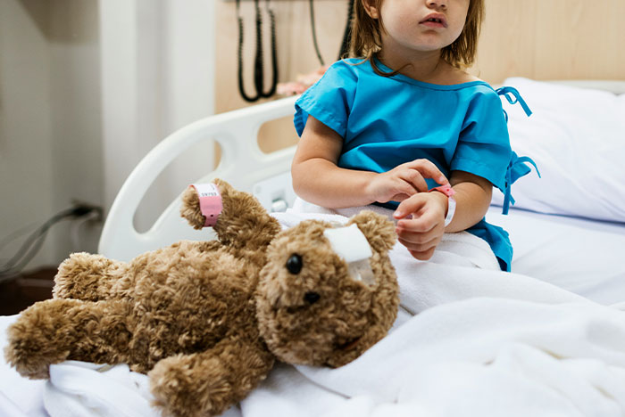 Child in hospital gown sitting on bed with a teddy bear, capturing moments when gut instinct was 100% correct.
